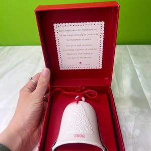 Pode incluir: Um ornamento de sino de porcelana branca com um design em relevo de uma árvore de Natal. O sino está em uma caixa vermelha com um cartão branco que diz "Os sinos têm sido uma parte importante da magia e da alegria do Natal por centenas de anos. Nosso sino de 2008 continua a tradição natalina de trazer felicidade e tocar no espírito da época."