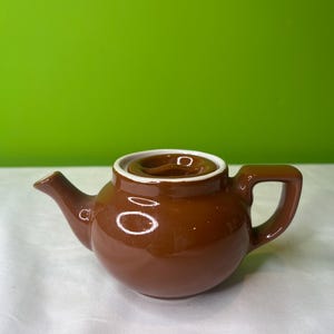 Puede incluir: Una tetera pequeña de cerámica marrón con tapa y asa blancas. La tetera está sobre una superficie blanca.