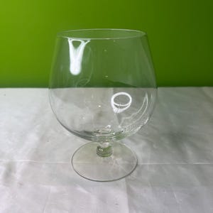 May include: A clear glass brandy snifter with a round bowl and a short stem. The glass is sitting on a white surface.