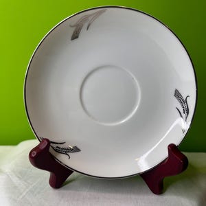 May include: White ceramic saucer with a silver rim and silver wheat design. The saucer is on a wooden stand.