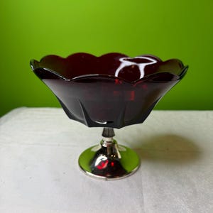 May include: A ruby-red glass pedestal bowl with a scalloped rim. The bowl sits on a silver-colored metal base. The bowl's design features a faceted exterior, adding to its vintage aesthetic. The background is a solid green.