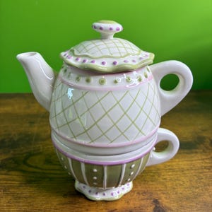 Peut inclure: Un service à thé en céramique composé d'une théière blanche à motif de treillis vert clair et accents roses, empilée sur une tasse assortie. Le couvercle de la théière présente un bord festonné à pois violets et la tasse des rayures verticales.