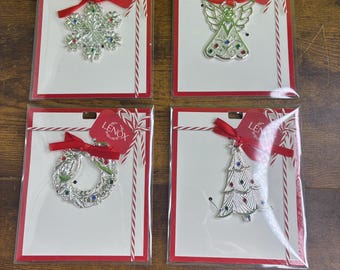 May include: Four silver-toned metal ornaments by Lenox, each packaged on a white card with a red border and red ribbon. The set includes a snowflake, an angel, a wreath, and a Christmas tree, all accented with small red, blue, and green gems.