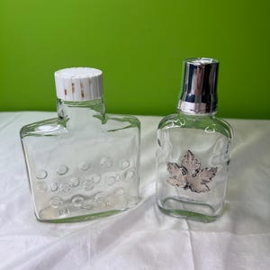 Puede incluir: Dos botellas de vidrio transparente. La botella de la izquierda tiene una forma rectangular ancha con una tapa blanca estriada y hendiduras circulares. La botella de la derecha es más estrecha con una tapa metálica plateada y una hoja de arce.