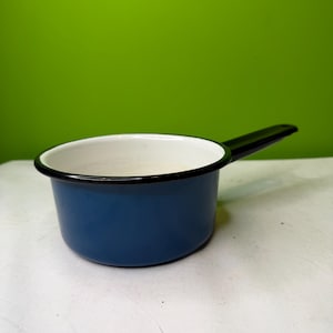 May include: A blue enamel saucepan with a black handle and rim. The interior is white. The saucepan is sitting on a white surface with a green background.