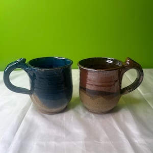 May include: Two handmade ceramic mugs. One is dark teal with a tan base, the other is brown with tan and brown bands. Both mugs have unique handles with a decorative flourish.
