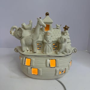 Könnte beinhalten: Cremefarbene Keramik-Noah-Arche-Figur mit goldenen Akzenten. Die Arche zeigt ein Haus mit einer Weihnachtsmannfigur, einem Weihnachtsbaum und verschiedenen Tieren. Die Arche hat beleuchtete Fenster und eine goldene Spitze.