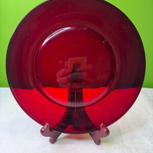 Könnte beinhalten: Eine tiefrote Glasplatte mit glänzendem Finish. Die Platte ist rund mit einem leicht erhöhten Rand und wird auf einem Holzständer präsentiert. Die Farbe ist durchgehend, ohne sichtbare Muster. Durchmesser ca. 25 cm.