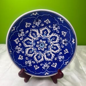 Puede incluir: Un plato decorativo redondo azul y blanco con un diseño floral. El plato se exhibe en un pequeño soporte de madera marrón oscuro. El diseño presenta un motivo floral central rodeado de elementos florales más pequeños.