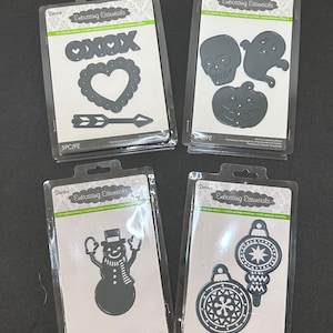 May include: Four packs of metal die-cut stencils for crafting. The stencils are packaged in clear plastic with black and white printed labels. The stencils include designs for hearts, arrows, skulls, ghosts, pumpkins, snowmen, and ornaments.