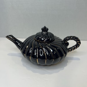 May include: A black ceramic teapot with a ribbed design and gold accents. The teapot has a curved spout, a looped handle, and a lid with a small crown-shaped finial. The teapot is on a white surface.