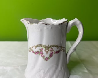 Puede incluir: Crema de cerámica blanca con un borde festoneado y un diseño floral en rosa y verde. La crema tiene un asa y una superficie texturizada.