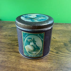 Może przedstawiać: Puszka Coca-Cola w stylu vintage z pokrywką. Puszka przedstawia kobietę w kapeluszu i ma ciemnobrązowy, żebrowany wzór. Zielona etykieta głosi "Delicious" i "Refreshing", z logo Coca-Cola. Puszka ma kształt cylindryczny.