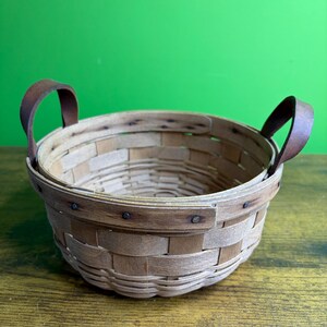 Puede incluir: Una cesta redonda tejida con tiras de madera de color marrón claro y dos asas de cuero marrón oscuro fijadas a los lados con remaches metálicos. La cesta presenta una estructura de borde superpuesto y una base circular de madera natural.