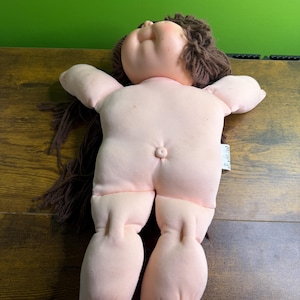Puede incluir: Una muñeca vintage con un cuerpo y extremidades rosas y suaves. La muñeca tiene una cara redonda con los ojos cerrados, mejillas rosadas y cabello de hilo marrón. Los brazos de la muñeca están extendidos y sus piernas están ligeramente dobladas.