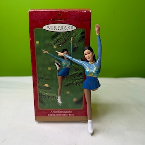 Peut inclure: Figurine d'ornement Kristi Yamaguchi Keepsake, représentant une patineuse artistique en costume bleu et or, les bras levés. L'ornement est devant une boîte rouge avec les mots "Keepsake Ornament" et "Kristi Yamaguchi Décoration des Fêtes".