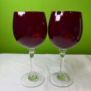 May include: A pair of wine glasses. Each glass has a deep red bowl and a clear stem and base. The glasses are on a white surface with a green background. The glasses are empty.