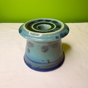 May include: A ceramic candle holder with a flared base and a wide, flat top. The piece is glazed in shades of blue and teal, with darker blue accents around the edges and a speckled pattern on the body. The top has a circular indentation.