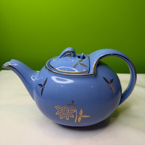 May include: A powder blue ceramic teapot with gold floral and leaf patterns. The teapot has a rounded shape, a spout, and a handle. Gold trim accents the lid and spout. The background is a solid green.