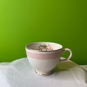 May include: A white teacup with a pink band and floral design. The cup has a gold rim and sits on a white surface.