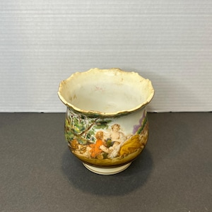 May include: A white ceramic planter with a scalloped rim and a painted scene of people in a garden. The planter has a brown base and a gold trim.