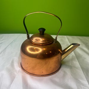 May include: A copper-colored tea kettle with a brass handle and spout. The kettle has a rounded body, a lid with a black knob, and a curved handle. The spout is angled, and the kettle sits on a white surface against a green background.