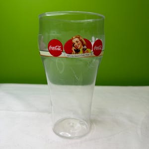 Könnte beinhalten: Ein durchsichtiges Coca-Cola-Glas aus Kunststoff mit Vintage-Design. Das Glas zeigt das Coca-Cola-Logo und ein Retro-Bild einer Frau, die einen Coke Float hält. Der Text auf dem Glas lautet: "Let's Go Have a Coke Float."