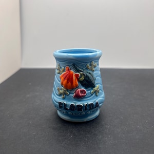 Puede incluir: Un pequeño vaso de chupito de cerámica azul claro con la palabra "FLORIDA" en letras negras. El vaso está decorado con diseños de vida marina naranja, negra y roja. El vaso tiene un diseño ondulado.