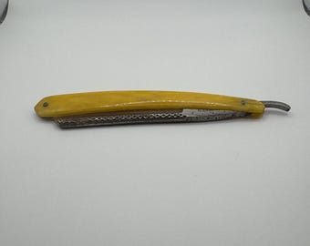 May include: A straight razor with a yellow handle and a silver blade. The blade is open, revealing its sharp edge. The handle is a light yellow color, and the razor appears to be vintage.