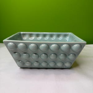 Puede incluir: Una jardinera cuadrada de cerámica azul claro con un patrón texturizado en forma de burbujas. La jardinera tiene una forma ligeramente acampanada y está sobre un fondo verde. Adecuada para uso en interiores o exteriores.