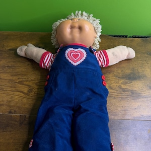Könnte beinhalten: Eine Vintage-Puppe mit gebräuntem Gesicht, blondem Garnhaar und ausgestreckten Armen. Die Puppe trägt eine blaue Cord-Latzhose mit roten herzförmigen Knöpfen und einer Herzapplikation. Die Ärmel sind rot-weiß gestreift.