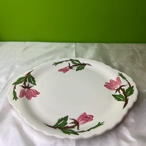 May include: White oval serving platter with a scalloped edge. The platter is decorated with pink floral designs and green leaves on brown stems. The background is a solid green.