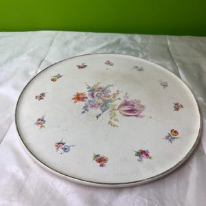 Może przedstawiać: Okrągła, biała ceramiczna taca do serwowania z kwiatowym wzorem. Taca ma centralny bukiet kolorowych kwiatów i mniejsze skupiska kwiatowe wokół krawędzi. Srebrna obwódka otacza tacę.