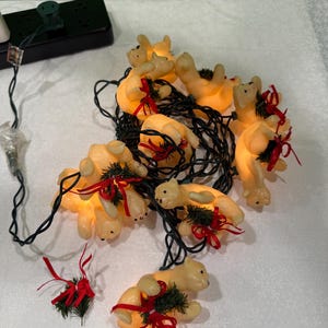 Könnte beinhalten: Eine Lichterkette mit beleuchteten Eisbärenfiguren. Jeder Bär ist cremefarben und mit einem roten Band und einem kleinen grünen Zweig geschmückt. Die Lichter sind an einem schwarzen Draht aufgereiht.