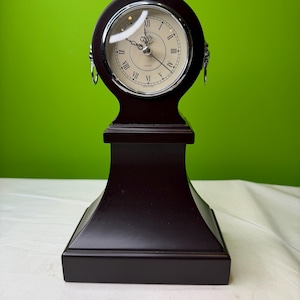 May include: A dark brown wooden quartz clock with a cream-colored face and Roman numerals. The clock has a circular face with a silver-colored bezel and decorative metal accents. The clock sits on a rectangular base.