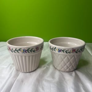 May include: Two white ceramic flower pots with a floral design around the top. One pot has a ribbed texture and the other has a diamond pattern.