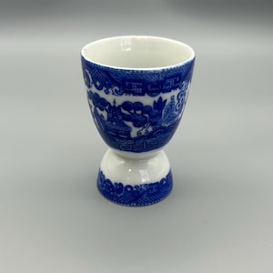 May include: A white and blue ceramic egg cup with a detailed Willow pattern design. The cup has a rounded top and a pedestal base. The blue pattern features a landscape scene. The egg cup is approximately 4 inches tall.