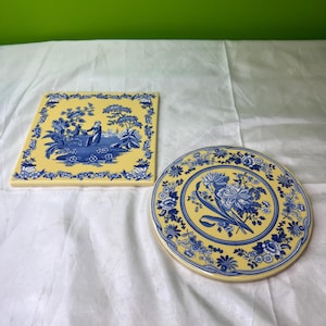 以下が含まれることがあります： 青と黄色の花柄のセラミック2点。1つは風景画の正方形、もう1つは花のブーケの円形です。どちらも黄色い背景に青い花の縁取りがあります。