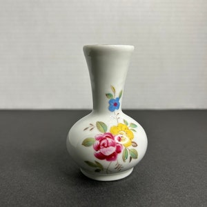 Puede incluir: Un pequeño jarrón de cerámica blanca con un diseño floral. El jarrón tiene un cuerpo redondo y un cuello estrecho. Las flores están pintadas en tonos de rosa, amarillo, azul y verde.