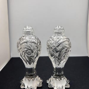 May include: Two clear, cut-glass salt and pepper shakers. Each shaker has a starburst pattern on the body and a faceted base. The lids are also cut glass and have a ridged design. The shakers are displayed on a dark surface.