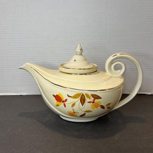 May include: A vintage teapot with a cream-colored body and a gold trim. The teapot has a floral design with orange, yellow, and brown leaves. The teapot has a unique genie lamp shape.