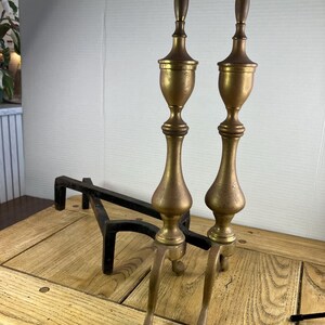 May include: Two antique brass and bronze fireplace andirons with a black iron grate. The andirons have a tall, ornate design with a bulbous midsection and a pointed top. The grate is rectangular with a curved front.