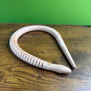 May include: A light-coloured wooden snake-shaped object with a segmented body and a pointed tail. The snake is lying on a wooden surface with a green background.