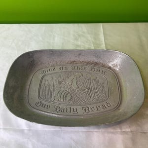 May include: A silver bread plate with an embossed design of wheat stalks and a person kneeling in a field. The plate reads "Give us this day our daily bread."