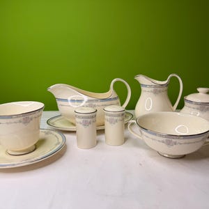 May include: A cream-colored porcelain tea set with blue and floral accents. The set includes a teacup and saucer, a gravy boat, a creamer, a sugar bowl, a soup bowl, and salt and pepper shakers.