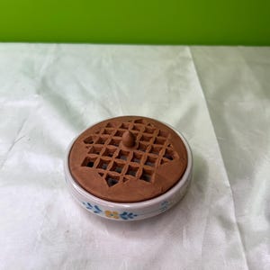 May include: A ceramic trinket dish with a brown, waffle-patterned lid. The dish has a white base with blue and yellow floral designs. The lid has a small knob for easy removal.