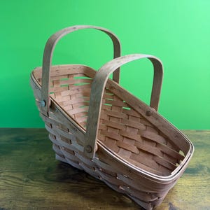 May include: A light brown woven wood basket featuring a rectangular base and two arched handles. The basket is constructed with thin, overlapping wooden strips in a lattice pattern, secured with small metal rivets at the handle attachment points.