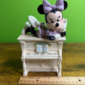 Peut inclure: Une figurine en c&eacute;ramique de Minnie Mouse portant une robe et un n&oelig;ud &agrave; pois lavande, pos&eacute;e sur un piano droit couleur cr&egrave;me avec un livre de musique. Une r&egrave;gle en bois plac&eacute;e devant montre que la base du piano mesure environ 14 cm de large.
