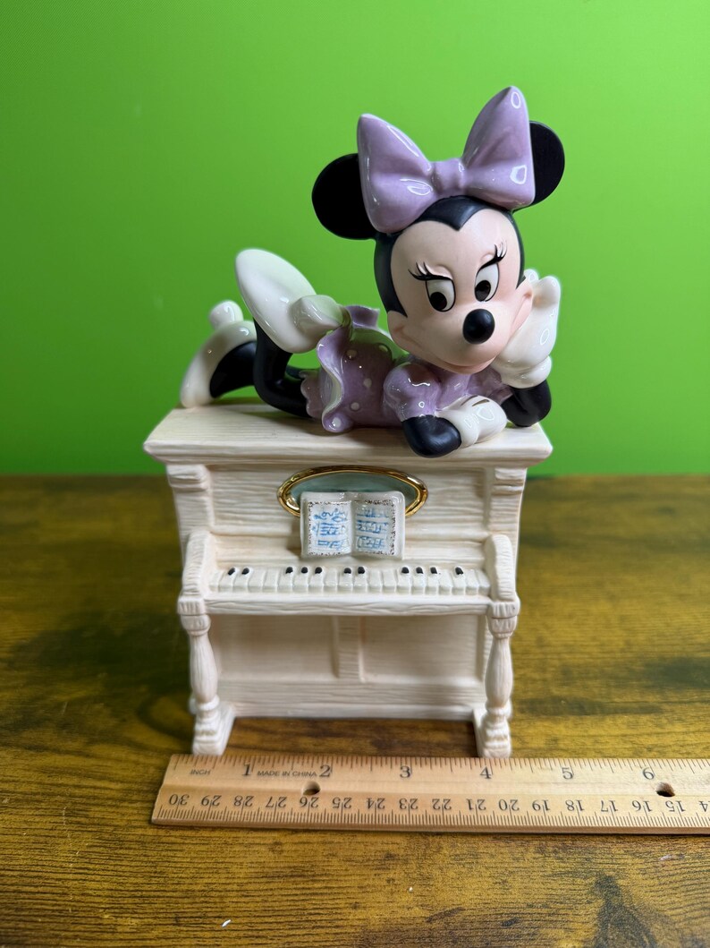 Peut inclure: Une figurine en c&eacute;ramique de Minnie Mouse portant une robe et un n&oelig;ud &agrave; pois lavande, pos&eacute;e sur un piano droit couleur cr&egrave;me avec un livre de musique. Une r&egrave;gle en bois plac&eacute;e devant montre que la base du piano mesure environ 14 cm de large.