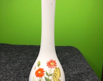 May include: A white ceramic vase with a floral design featuring red and yellow flowers, green leaves, and a yellow butterfly. The vase has a narrow neck and a rounded base.